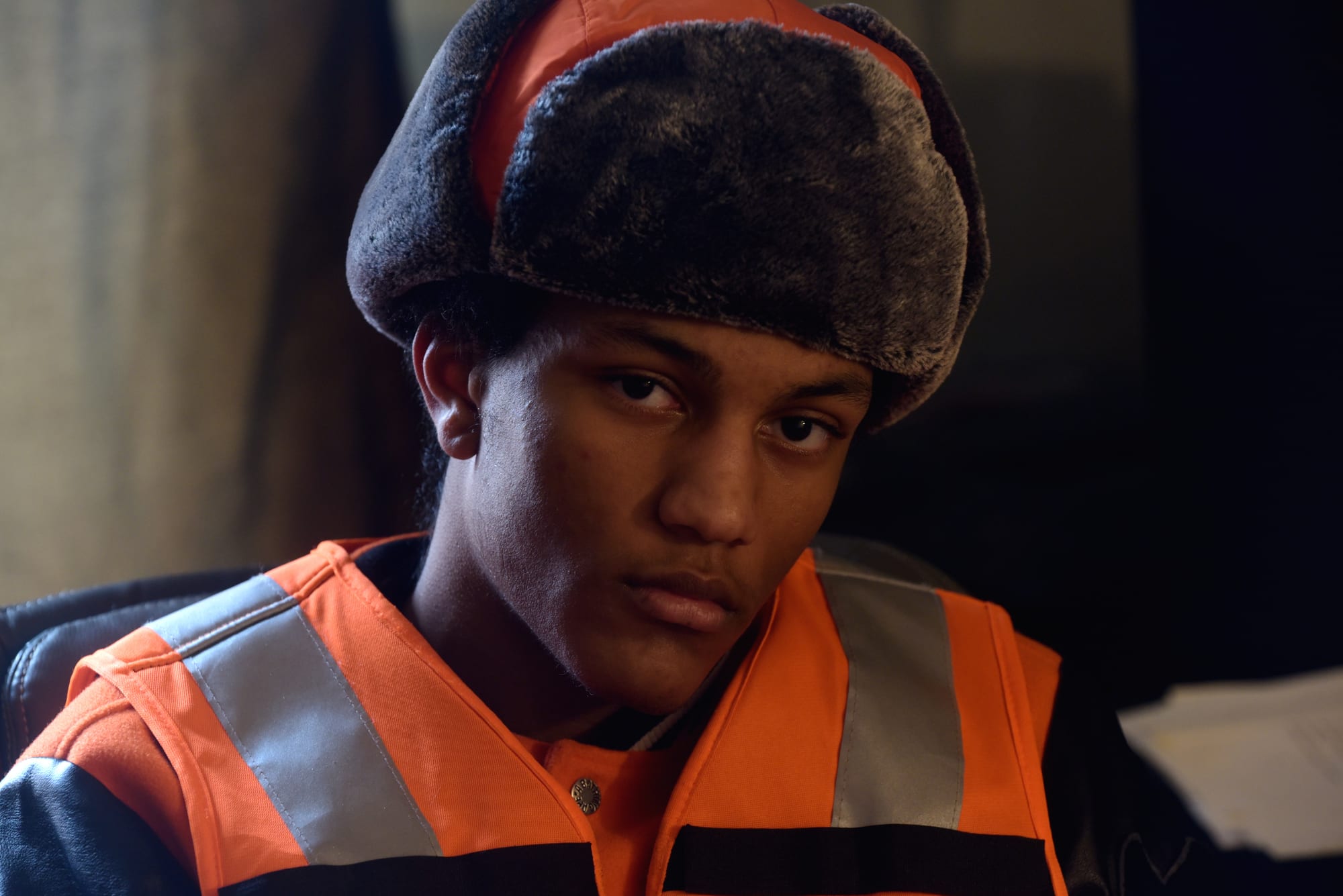 Young man in a faux-fur trapper hat and reflective orange vest over a leather jacket looks at the camera in warm low light. (file photograph)
